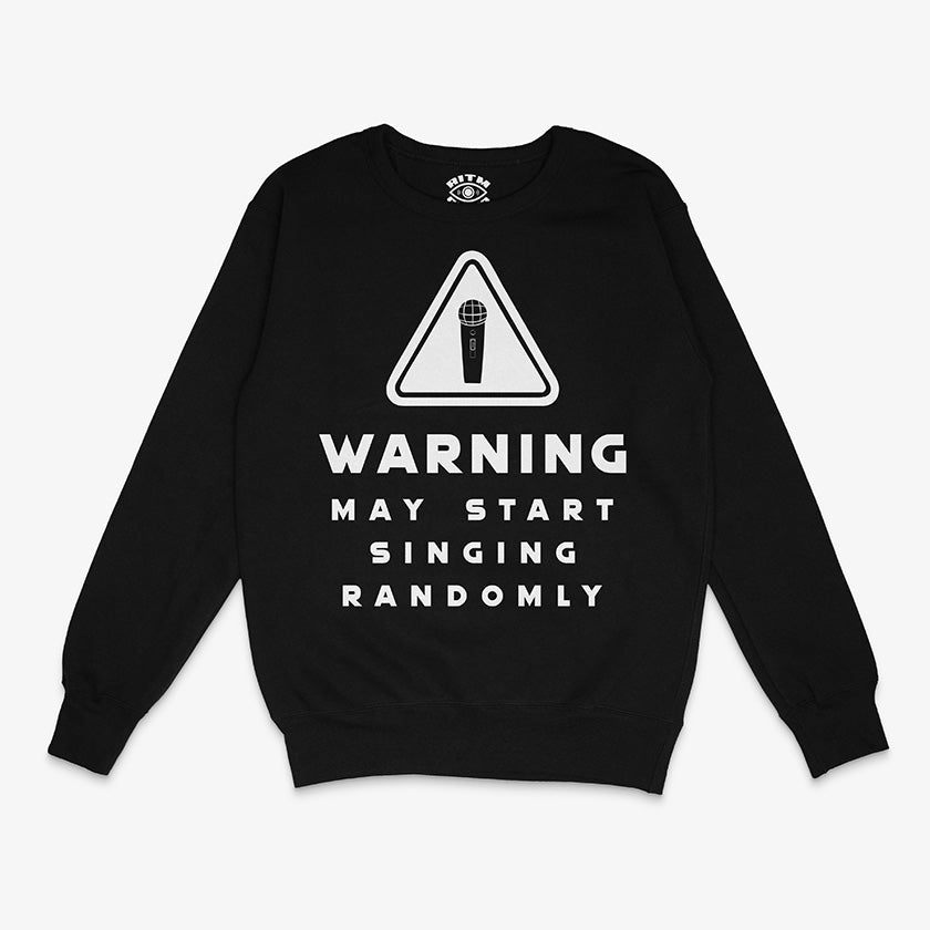 Polerón de cuello redondo de color negro. Diseño frontal con un icono de advertencia (triángulo) y un micrófono, con el texto en inglés "WARNING MAY START SINGING RANDOMLY"